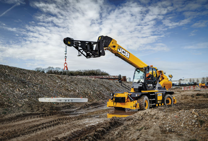 A JCB 555-210R Rotating Telehandler driving through a muddy site with it carrying concrete on a crane attachment