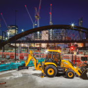 JCB 3CX Plus excavating with rear arm at night in front of a bridge