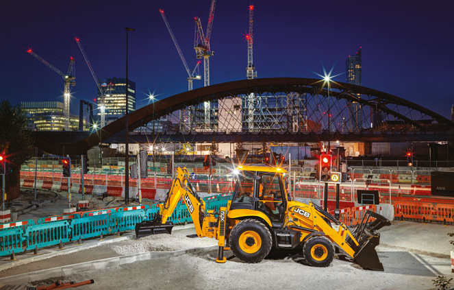 JCB 3CX Plus excavating with rear arm at night in front of a bridge
